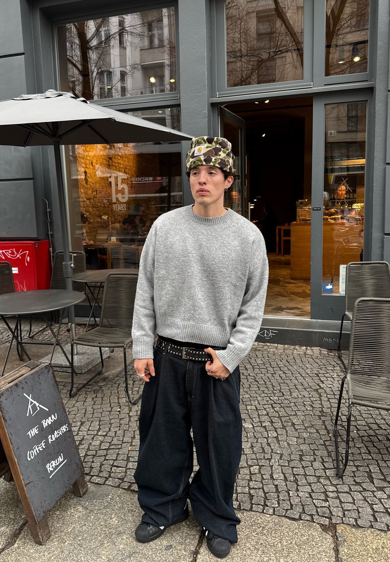 Young person wearing a grey sweater and camo hat stands outside a café with outdoor tables and a sign reading "The Barn Coffee Roasters Berlin."