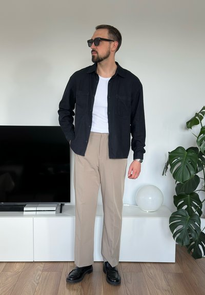 Camisa negra de botones sobre una camiseta blanca, pantalones beiges y zapatos negros. Diseño minimalista, textura suave, llevada en interiores por una persona con barba.