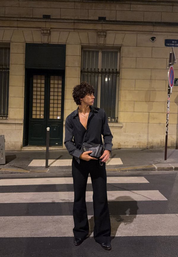 Jeune personne aux cheveux bouclés portant une tenue noire, debout sur un passage pour piétons en ville la nuit, tenant un petit sac et regardant de côté.