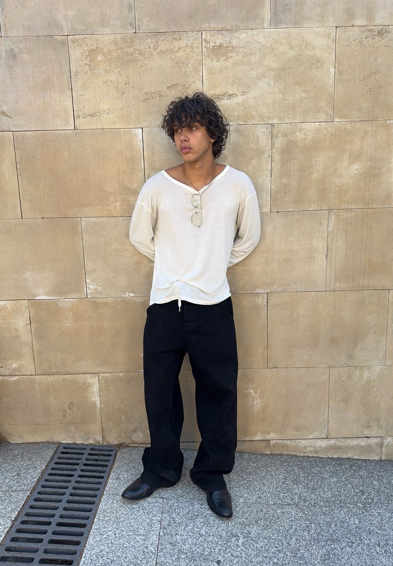 Young person with curly hair stands against beige stone wall, wearing white shirt, black pants, black shoes, and glasses hanging from shirt collar.