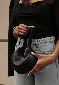 Črna usnjena torbica z edinstveno geometrijsko obliko, okroglo zgoraj in kratkimi ročaji, z gladko teksturo in minimalno okovje.