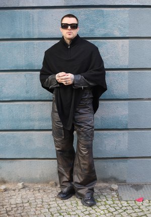 Poncho negro sobre una camiseta gris de manga larga, combinado con pantalones de cuero marrón holgados y zapatos negros. Las gafas de sol acentúan el conjunto.