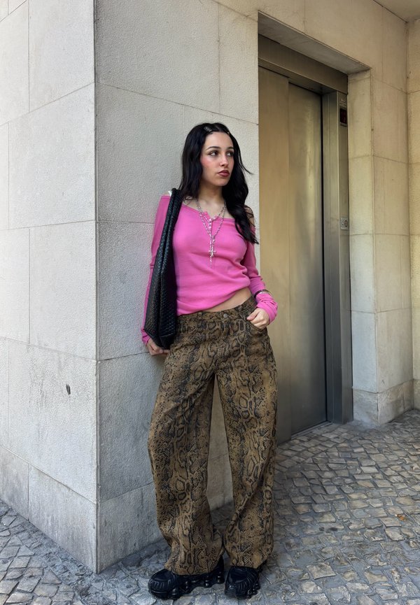 Femeie cu păr negru și lung, purtând un top roz cu mânecă lungă, pantaloni largi cu imprimeu de șarpe, pantofi negri și care poartă o geantă neagră lângă ușa unui lift.