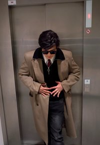 Homme en manteau beige et lunettes de soleil, se tenant le ventre, debout devant des portes d'ascenseur fermées affichant le numéro de l'étage un.