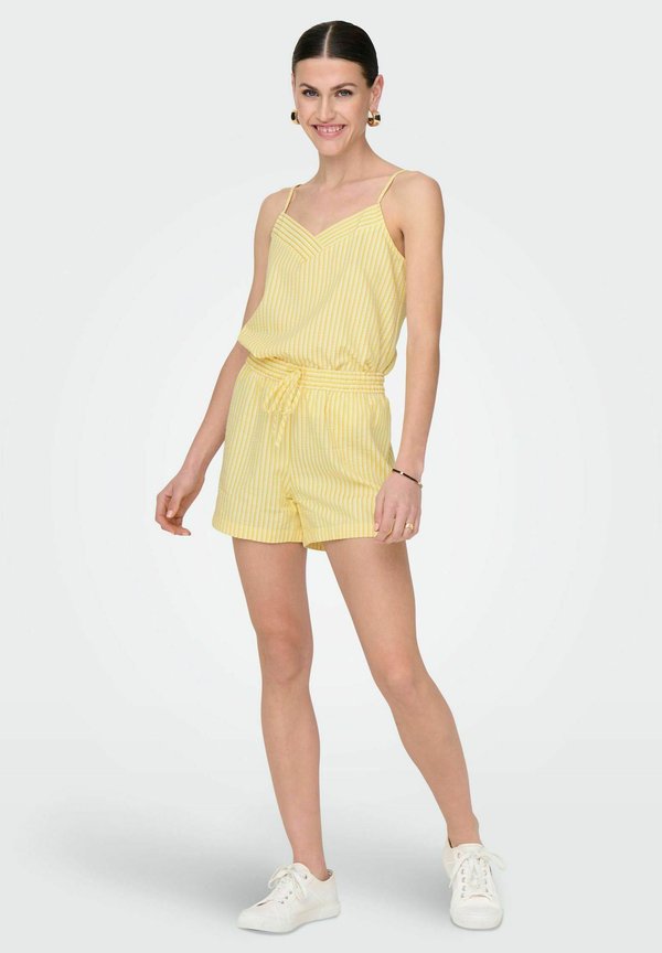 Combinaison à rayures jaunes avec des bretelles spaghetti et une taille cintrée. Associée à des baskets blanches, mettant en avant un look décontracté et estival.