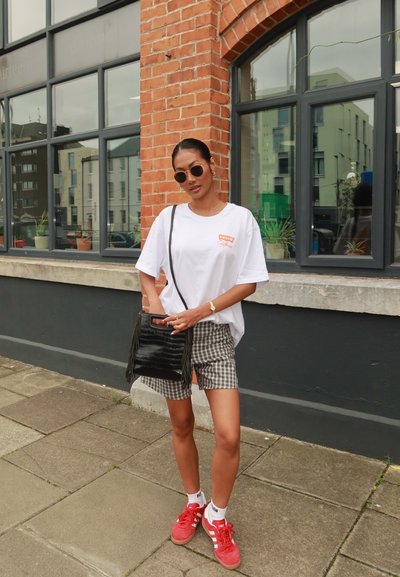 Camiseta blanca de gran tamaño con logo rojo, pantalones cortos de cuadros, bolso negro con flecos, zapatillas rojas y gafas de sol redondas contra una pared de ladrillo.