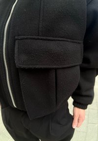 Μαύρο φούτερ με φερμουάρ μπροστά και μεγάλη, υφασμάτινη τσέπη στην αριστερή πλευρά. Διαθέτει άνετη εφαρμογή και λείο υλικό.