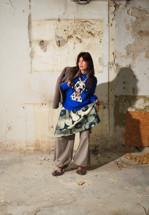 Mujer con suéter azul con estampado de perros, falda floral sobre pantalones de cuadros, sosteniendo un abrigo sobre su hombro en una habitación desgastada y texturizada.
