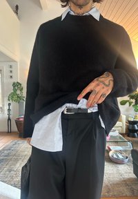 Sort strikket trøje over en hvid skjorte med krave, kombineret med sorte bukser. Bemærkelsesværdige tatoveringer på hænderne og en sølvfarvet bæltespænde.