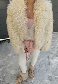 Personne portant un manteau crème duveteux, des leggings blancs, des bottes beiges en fourrure, tenant un sac à main matelassé rose, debout sur un sol recouvert de neige.
