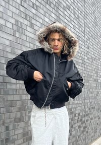 Czarna kurtka typu bomber z kapturem wyłożonym futerkiem, zapinana na zamek, o luźnym kroju. Noszona na szarych spodniach dresowych na tle teksturowanej ceglanej ściany.