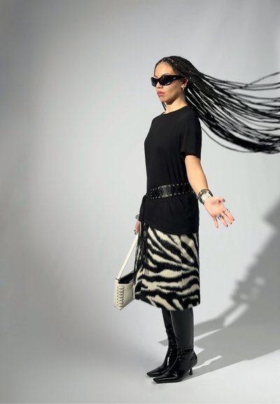 Mujer con cabello largo trenzado que gira de lado, lleva gafas de sol negras, camisa negra, falda con estampado de cebra, botas negras y sostiene un bolso blanco.