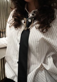 Femme portant un chemisier blanc à fines rayures avec des boutons de manchette en perle et une cravate noire décorée de chaînes dorées ainsi que de breloques en forme de cœur et de nœud.