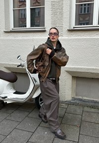 Barna bőrdzseki zöld kapucnis pulóver és fehér ing fölött, széles szürke nadrággal párosítva. Fekete csizmák és barna táska. Egy robogó közelben.