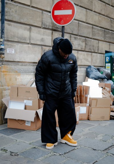 Chaqueta de plumas negra con un logo, pantalones negros oversized y botas de plataforma amarillas brillantes. El fondo incluye cajas de cartón y un cartel de prohibido el paso.