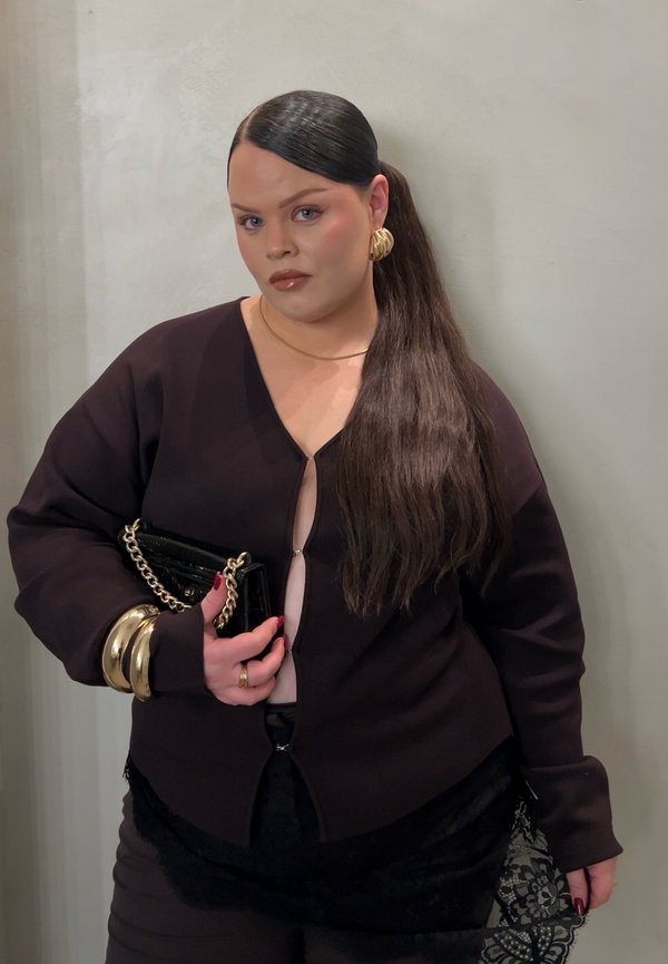 Top nero a maniche lunghe con scollo a V profondo e zip, abbinato a una gonna di pizzo nera. Orecchini a cerchio dorati e braccialetti, con una clutch nera in mano.