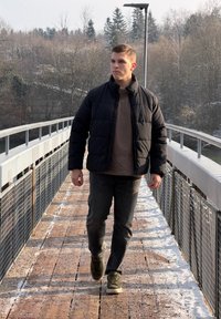 Fekete pufi dzseki, barna ing és sötét farmernadrág. Egy fémkorlátokkal szegélyezett fa hídon sétál, téli táj veszi körül.