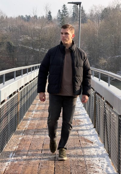 Chaqueta negra acolchada, camisa marrón y vaqueros oscuros. Caminando sobre un puente de madera con barandillas metálicas, rodeado de un paisaje invernal.