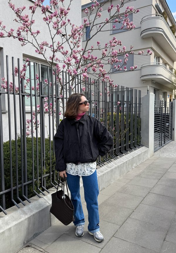 Femme portant des lunettes de soleil et une veste noire, tenant un sac, debout sur le trottoir près d'une clôture métallique noire avec un magnolia rose en fleurs.