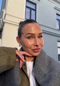 Chaqueta verde oliva con cuello de forro polar gris, camiseta blanca debajo, uñas naturales, pendientes de aro y piel suave, con un fondo claro.