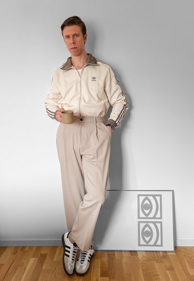 Chaqueta zip-up crema de Adidas con acentos marrones, pantalones beiges con pliegues y zapatillas blancas con rayas negras. Sosteniendo una taza beige.