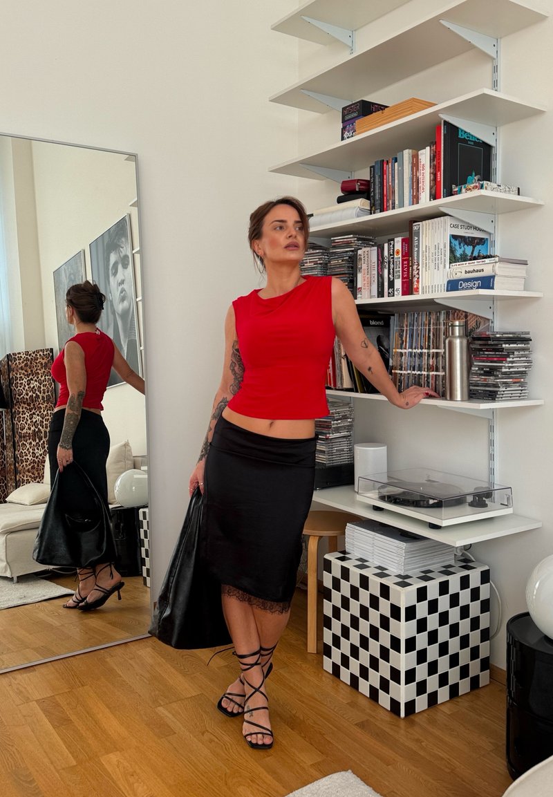 Kvinde med tatoveringer iført rød top og sort nederdel holder en sort taske og læner sig op ad en hylde nær bøger, CD'er og en pladespiller i et moderne rum.