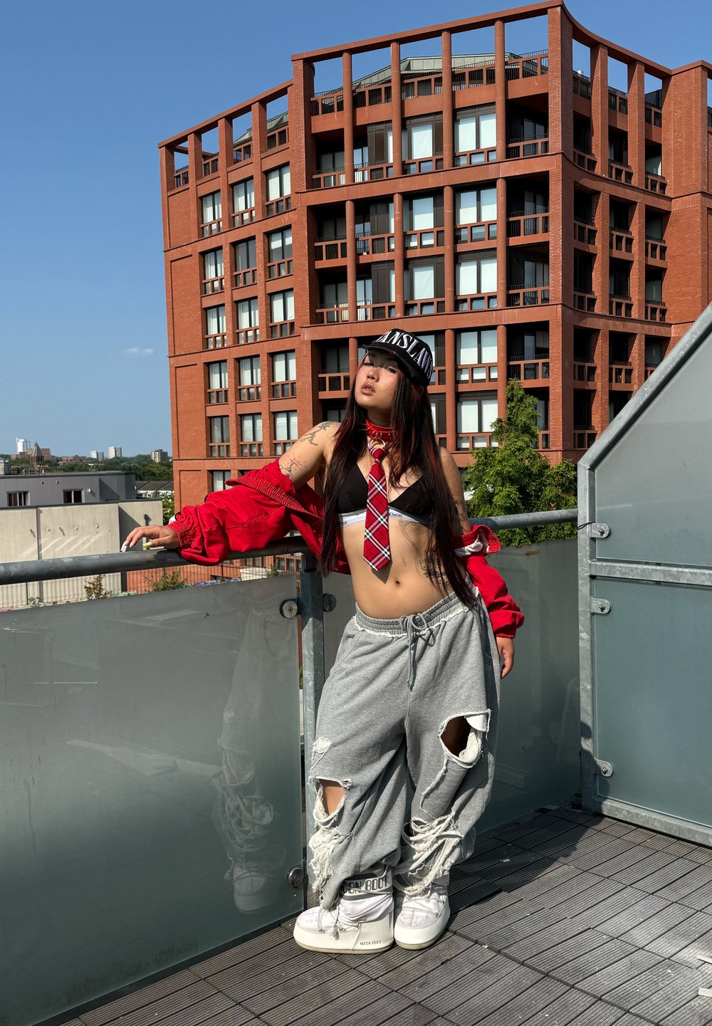 Modellen bär en röd oversized jacka, svart croptop, rutigt slips, grå mjukisbyxor med hål och vita högklackade skor framför en modern byggnadsbakgrund.