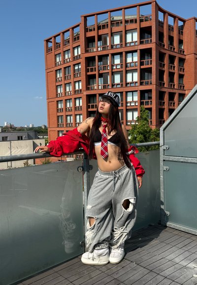 La modelo lleva una chaqueta roja oversized, un top corto negro, una corbata de cuadros, pantalones de chándal grises con desgarros y zapatillas altas blancas, todo ello frente a un moderno edificio.