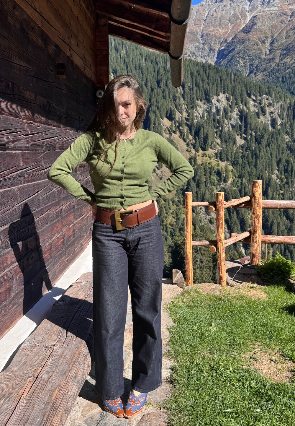 Grønn cardigan, mørk jeans, brunt belte med en firkantet spenne og mønstrede tresko. Står mot en trestruktur med naturskjønn bakgrunn.