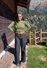 Grønn cardigan, mørk jeans, brunt belte med en firkantet spenne og mønstrede tresko. Står mot en trestruktur med naturskjønn bakgrunn.