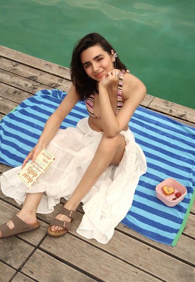 Top de bikini a rayas, falda blanca en capas, sandalias marrones. Toalla a rayas azul y verde, recipiente rosa con fruta, terraza de madera, agua verde.