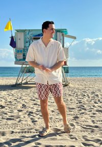 Άνδρας με λευκό κοντομάνικο πουκάμισο και κόκκινο σορτς με σχέδια που στέκεται σε αμμώδη παραλία κοντά σε πύργο ναυαγοσώστη με ωκεανό και ουρανό στο φόντο.