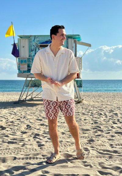 Hombre con camisa blanca de manga corta y pantalones cortos rojos con estampado, de pie en una playa de arena cerca de una torre de vigilancia con el océano y el cielo de fondo.