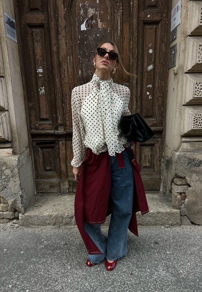 Femme portant des lunettes de soleil noires, chemisier blanc transparent à pois, jean à jambes larges, manteau bordeaux noué à la taille, chaussures rouges ouvertes à l’avant, tenant un sac noir.