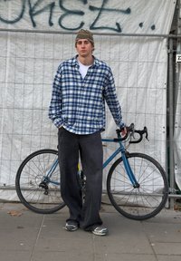 Une chemise à carreaux bleue, un pantalon ample foncé et des baskets grises portés par une personne debout à côté d'un vélo bleu avec des guidons abaissés.