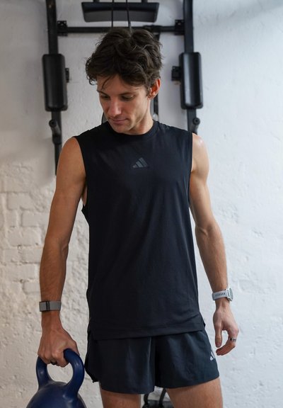 Hombre con camiseta sin mangas negra y pantalones cortos sosteniendo una pesa rusa azul en un gimnasio con equipo de entrenamiento al fondo.