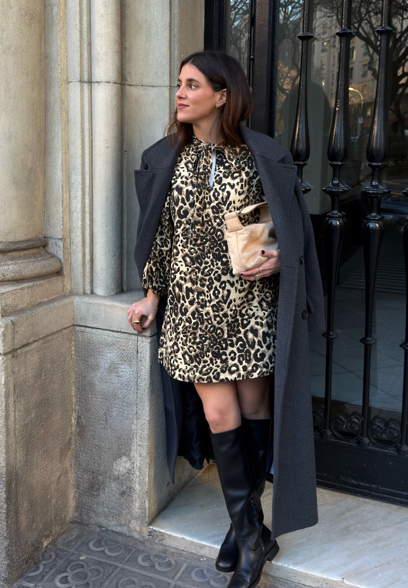 Femme en robe à imprimé léopard et manteau foncé appuyée contre un pilier en pierre, tenant un sac à main beige, portant des bottes noires montantes près d'une grille en fer noir.