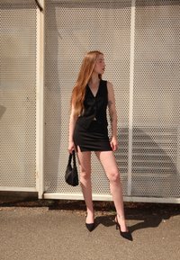 Débardeur noir avec des boutons associé à une mini-jupe noire avec des fentes sur les côtés. Le modèle tient un sac à main noir et porte des talons aiguilles.