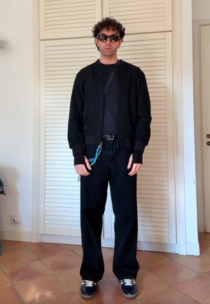 Chaqueta bomber negra con puños de canalé, combinada con una camiseta negra y jeans negros de pierna ancha. Las zapatillas tienen detalles y cordones blancos.