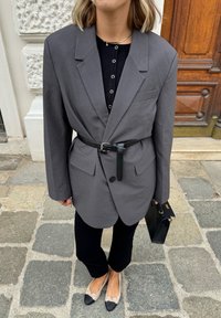Sacou gri supradimensionat cu o curea neagră, purtat cu un top negru, pantaloni negri și balerini bej cu vârfuri negre. Geantă de umăr neagră.
