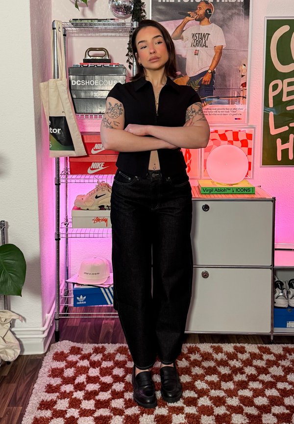 Jeune femme avec des tatouages sur les deux bras, les bras croisés, dans une pièce avec des baskets, des affiches et un éclairage rose.