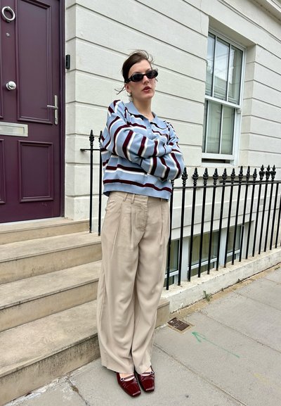 Suéter ligero a rayas en tonos azules y granates, combinado con pantalones beige holgados y zapatos rojos brillantes, de pie junto a un edificio blanco.