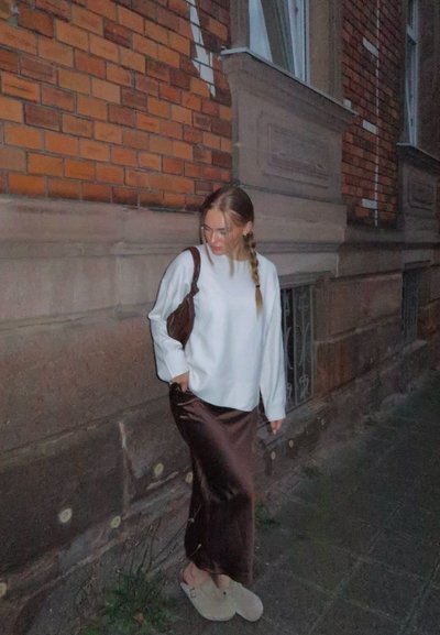 Joven mujer con trenzas que lleva un suéter blanco, una falda de satén marrón, zapatos beige tipo slip-on y un bolso de hombro marrón, caminando por una acera de noche.