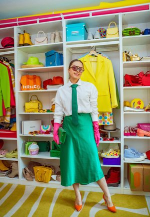 Mujer con falda verde, camiseta blanca, guantes rosas y tacones naranjas de pie frente a estantes blancos con zapatos, bolsos y ropa de colores.