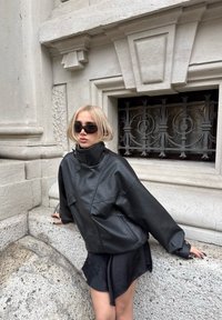 Schwarze Lederjacke mit übergroßem Design, hohem Kragen und zwei Vordertaschen, kombiniert mit einem schwarzen Rock und Sonnenbrille. Strukturierter Stein-Hintergrund.