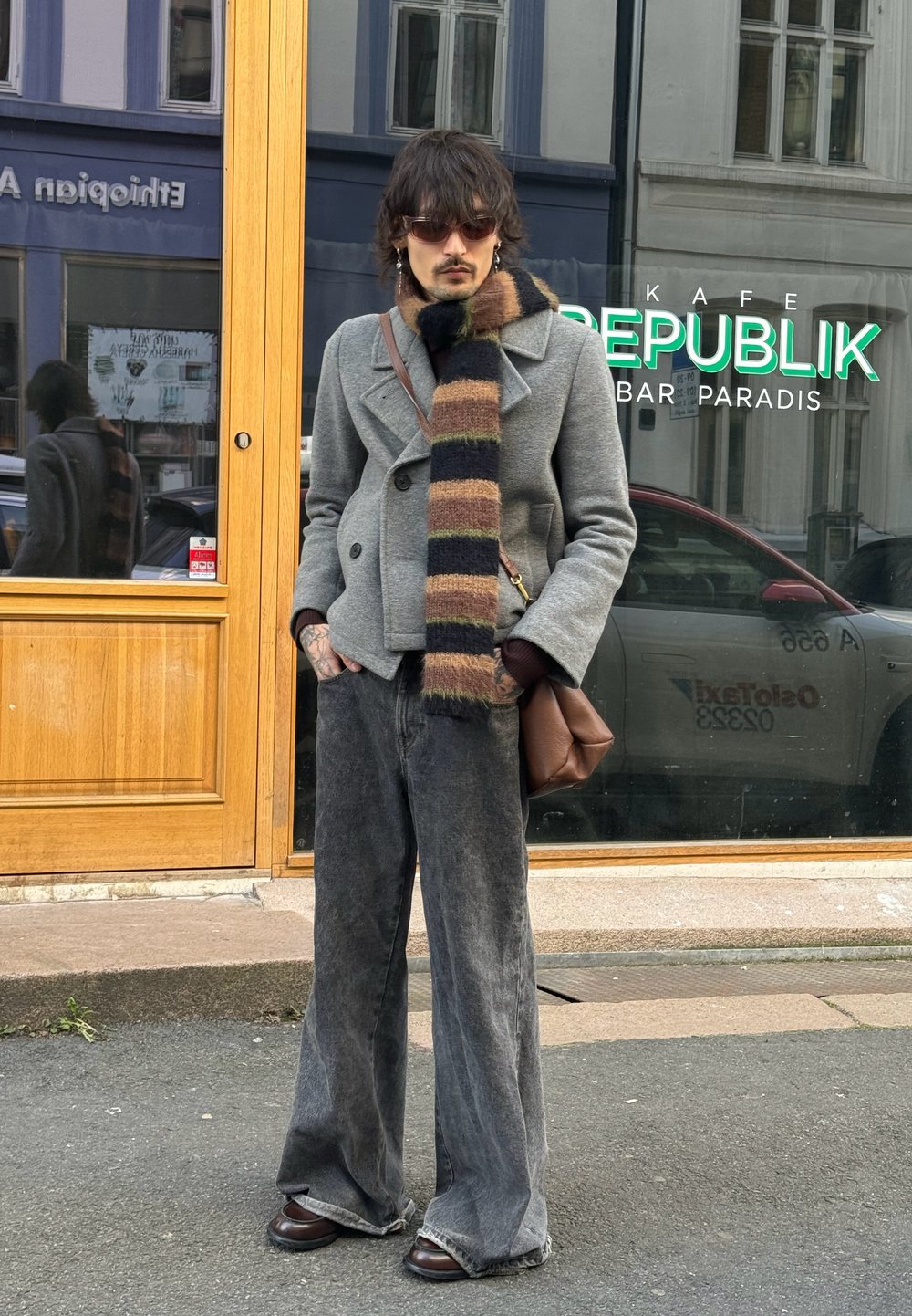 Manteau gris croisé, écharpe rayée noire et marron, jean large bleu foncé, chaussures en cuir marron et sac marron. Scène de rue avec fenêtre de café.