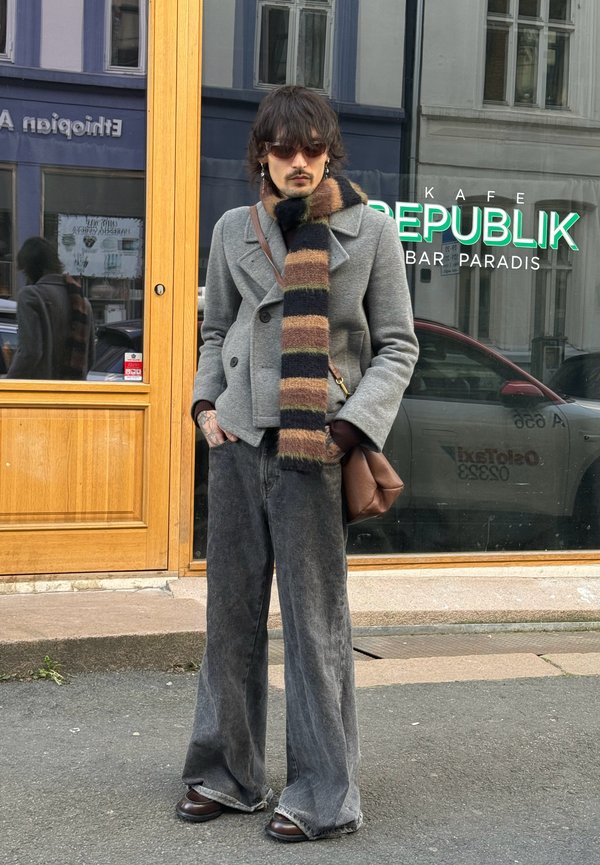 Gray double-breasted coat, black and brown striped scarf, wide-leg dark jeans, brown leather shoes, and a brown bag. Street setting with café window.