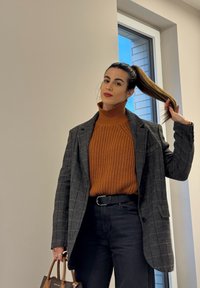 Sivi srajčni blazer s karo vzorcem, oranžen pleteni rolk, črne hlače, drži rjavo torbico. Ravne teksture z izrazitim pletenjem in krojenimi oblikami.