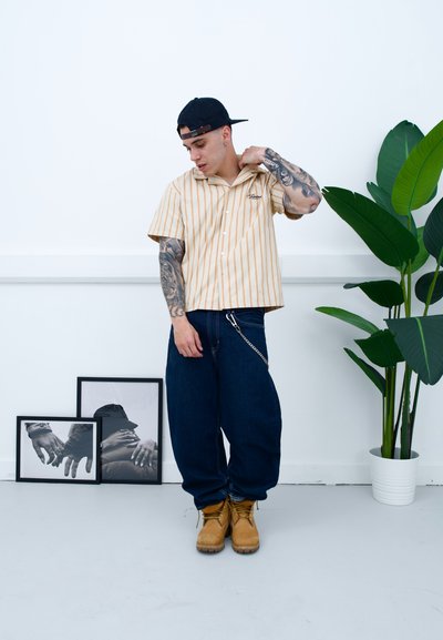 Camisa de manga corta a rayas en beige y blanco, combinada con unos vaqueros holgados de color azul oscuro y botas de trabajo en tono marrón. Incluye un accesorio de cadena y tatuajes.