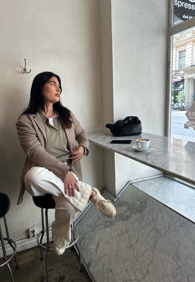 Blazer marrón claro sobre una camisa verde, pantalones anchos crema y zapatillas blancas. Mesa de mármol, bolso negro trenzado y taza de café cerca.